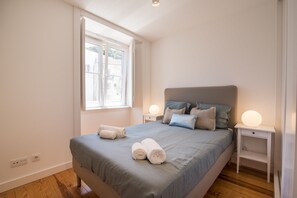 A neatly made bed with a grey bedspread and two rolled white towels, a bedside table with a lamp, and a window with a view of buildings. - Alfama Premium by Homing (Lisbon)