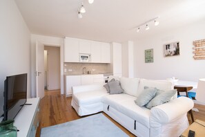 Una sala de estar moderna con un sofá blanco, un televisor de pantalla plana y una cocina con armarios blancos y microondas.