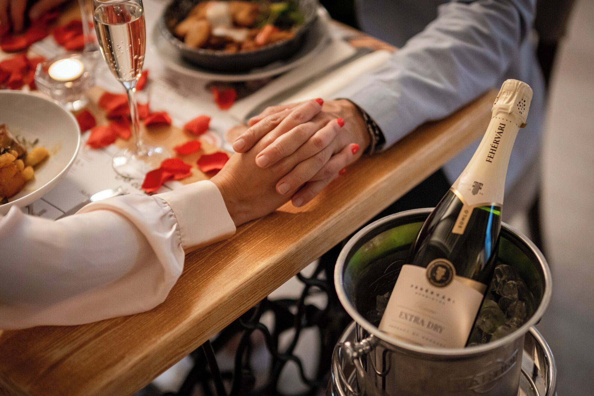 A person holding hands with another, a bottle of champagne in a bucket, and a plate of food on a table.