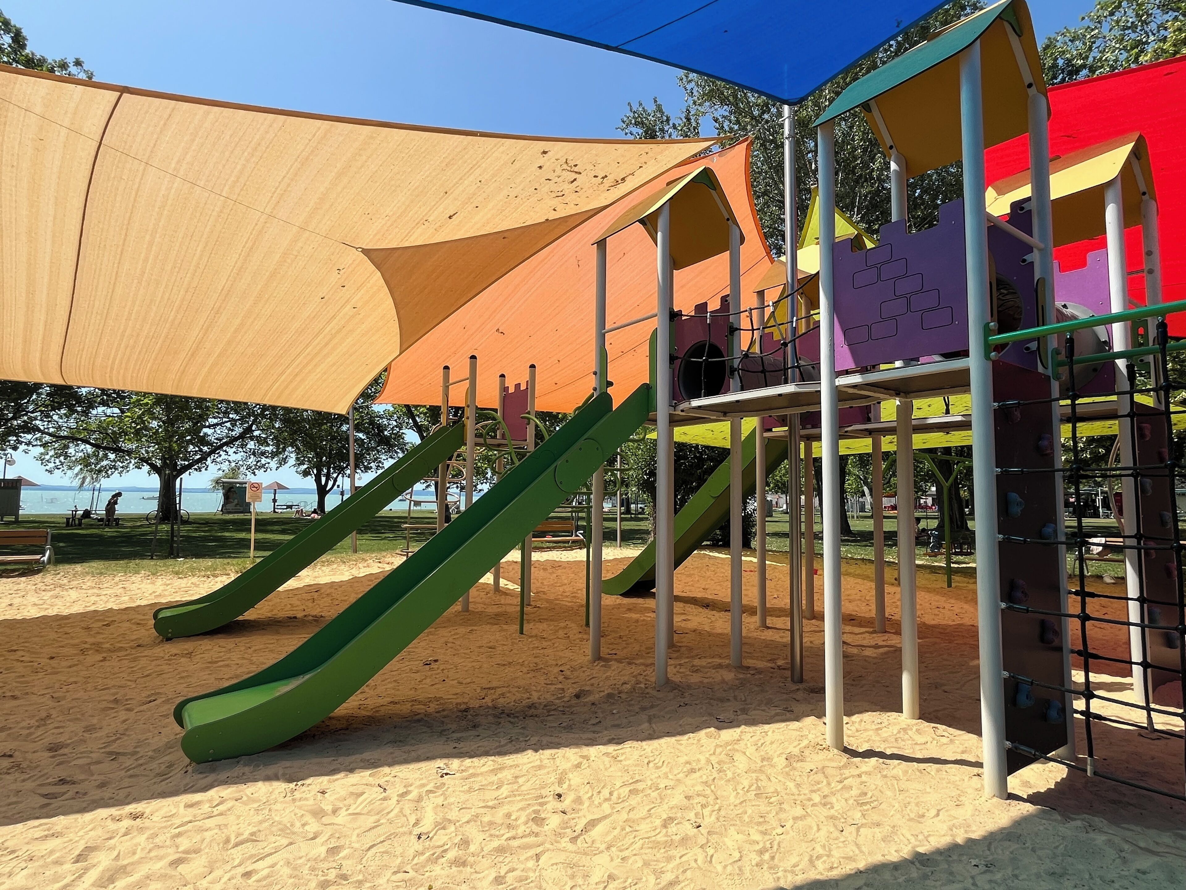 Un parque infantil con toboganes y estructuras para escalar, rodeado de arena y árboles.