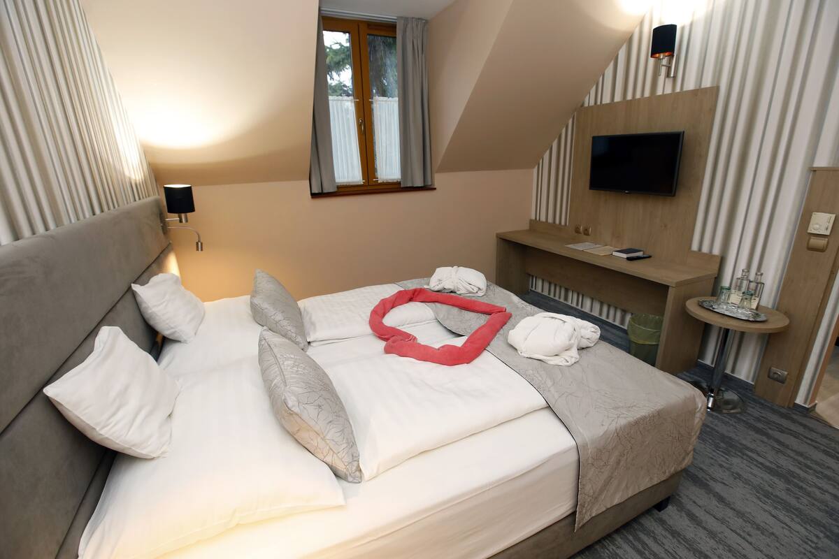 A hotel room with a bed, a TV mounted on the wall, a small table with a glass, and a window with curtains.