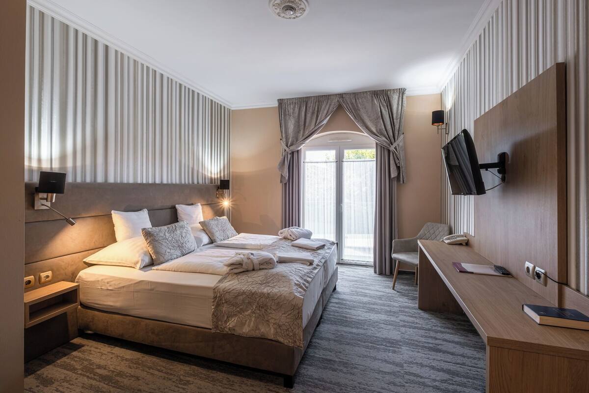 A hotel room with a large bed, bedside tables, a desk, a TV, and a window with curtains.