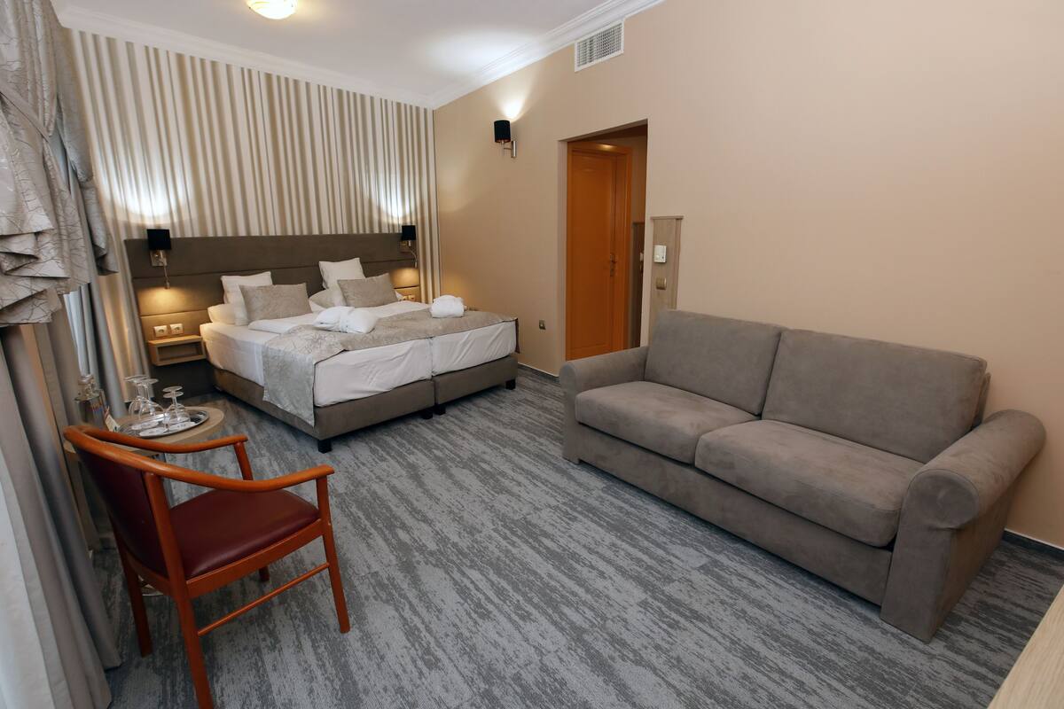 A hotel room with a bed, a sofa, a chair, and a small table with a glass set.