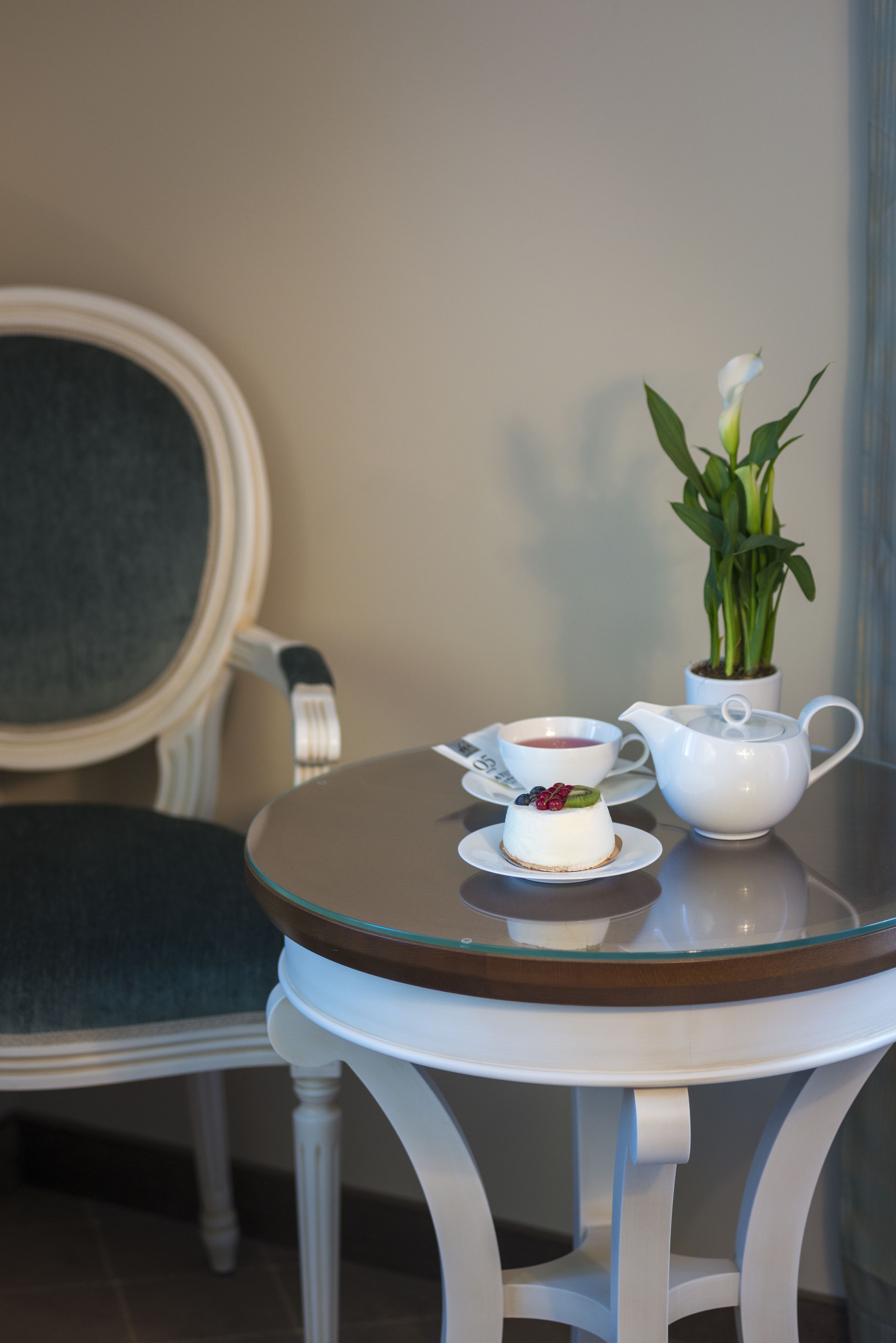 Una mesa con un calentador de agua, una taza y un postre sobre una superficie de vidrio, una silla y una planta en maceta.
