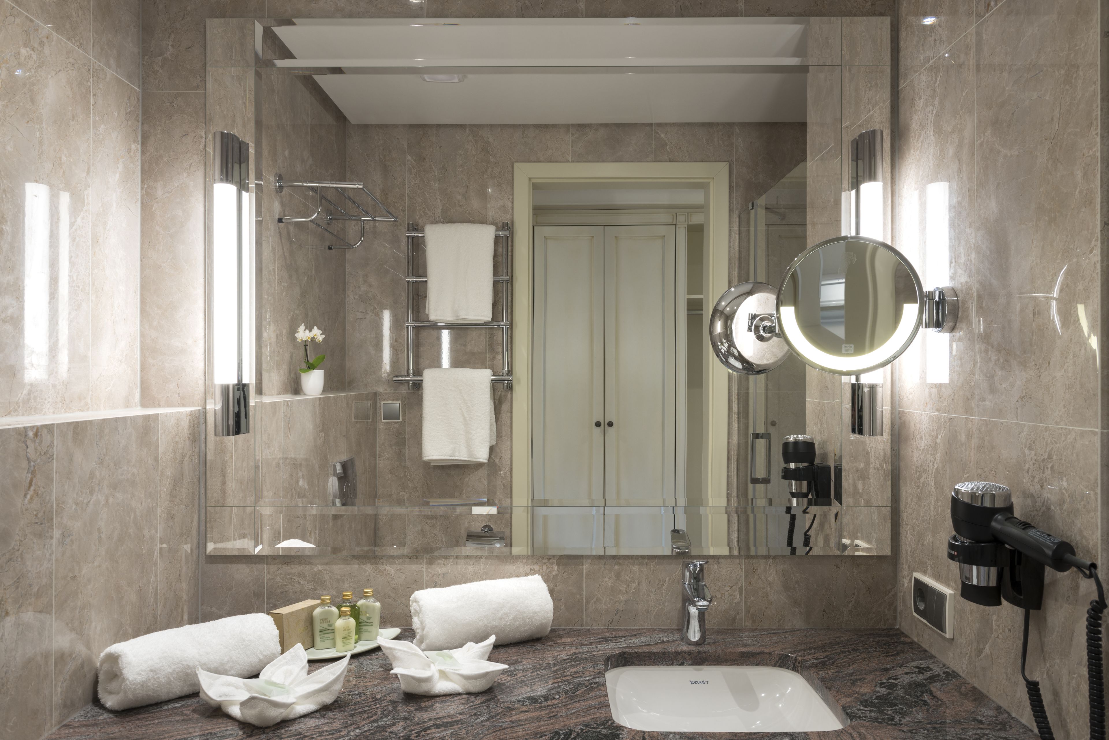 A modern bathroom with a marble sink, a large mirror, and a hairdryer mounted on the wall.