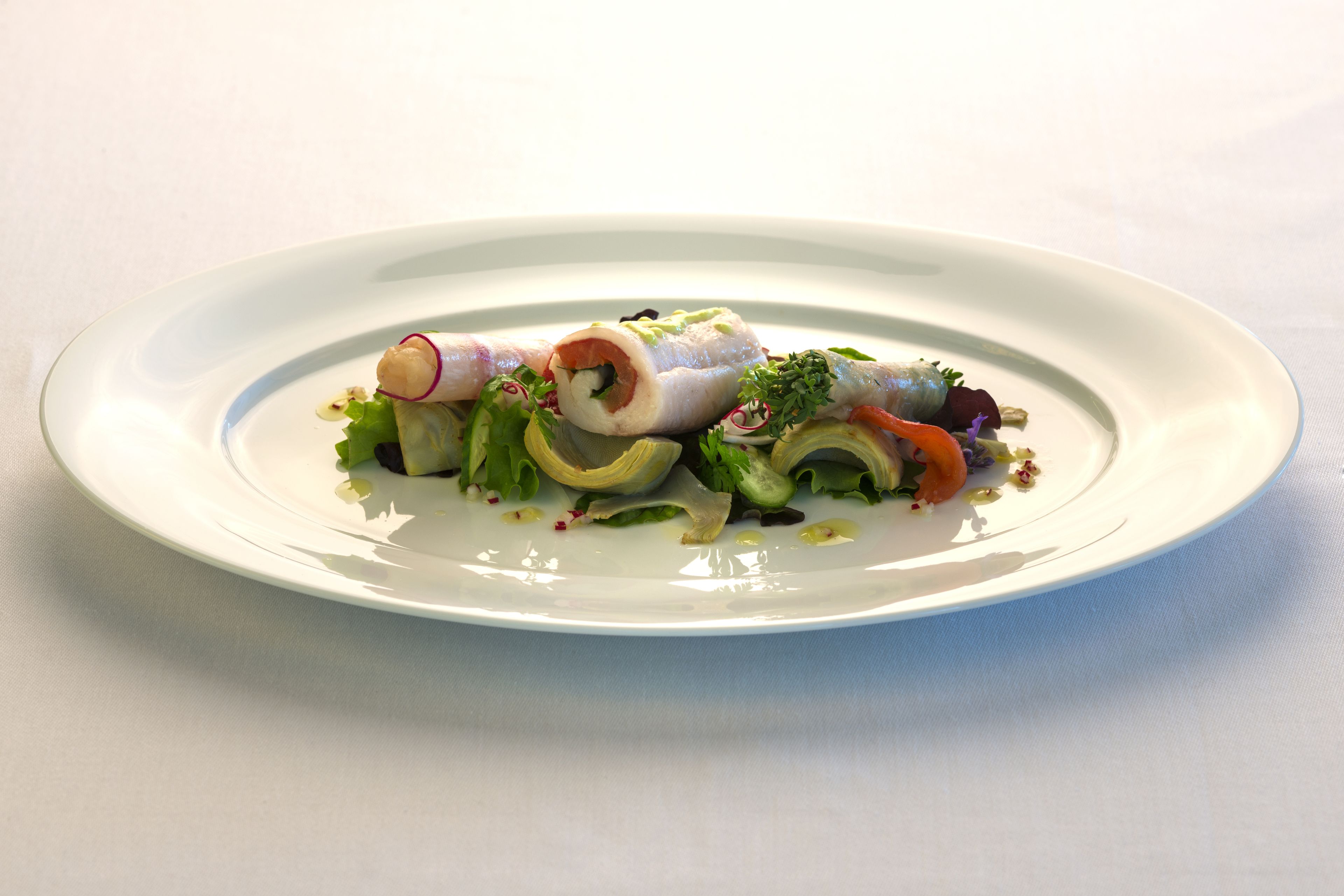 A white plate with a seafood dish, including rolled fish and vegetables, on a white tablecloth.