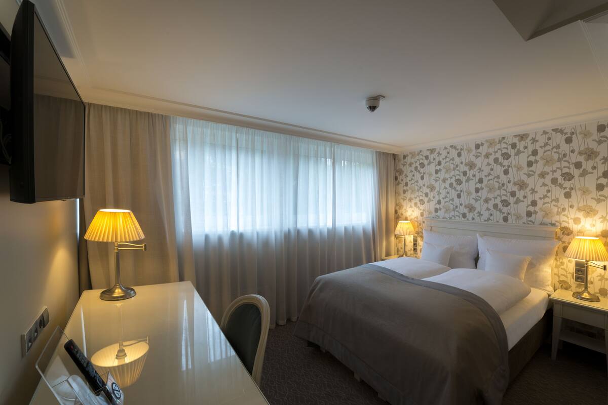Ein Hotelzimmer mit einem Bett, zwei Nachttischlampen, einem Schreibtisch mit Lampe, einem Sessel und einem Fernseher an der Wand.