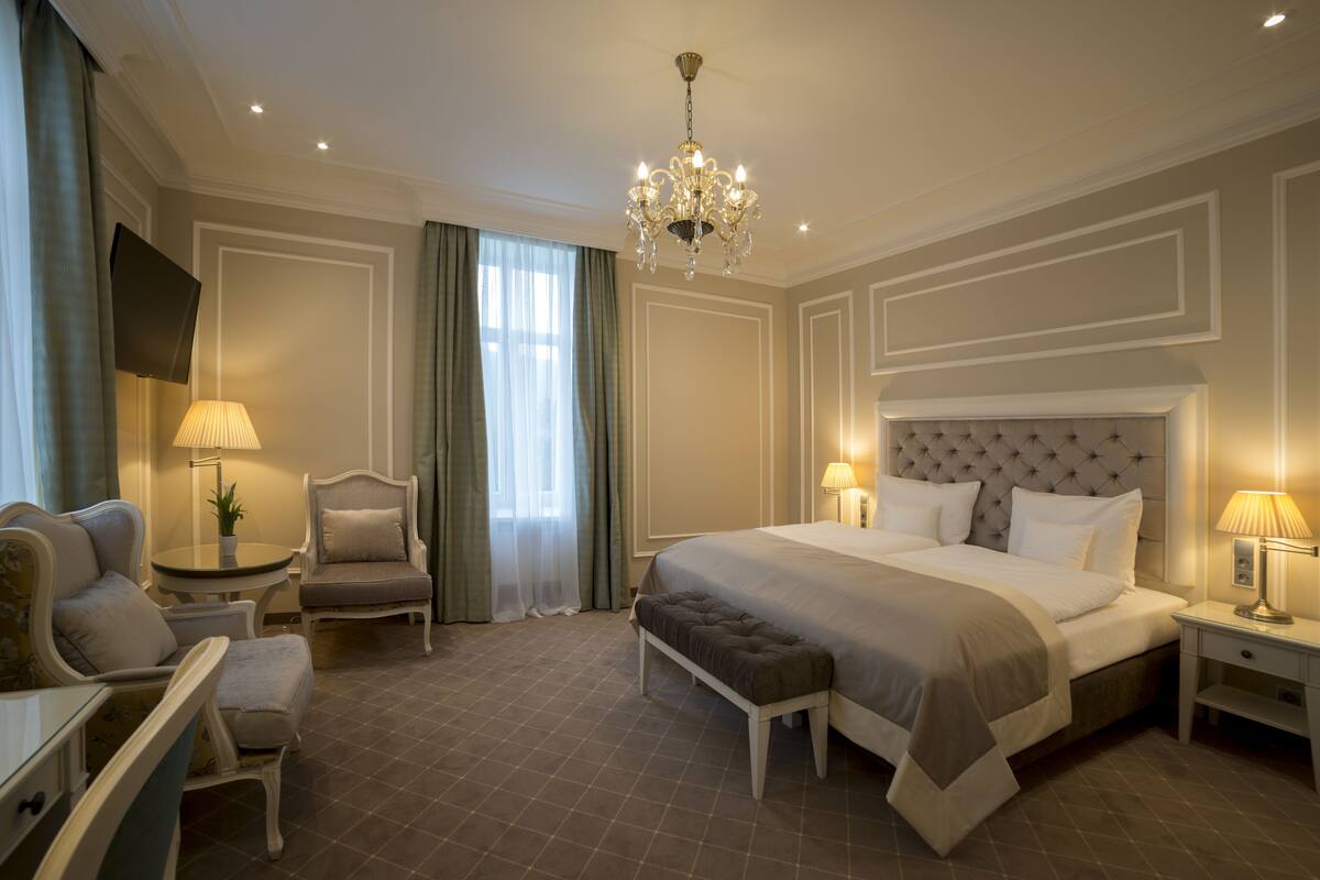 Ein Hotelzimmer mit einem großen Bett, einer Sitzecke mit Stuhl und Tisch, einer Bank und einem an der Wand befestigten Fernseher.