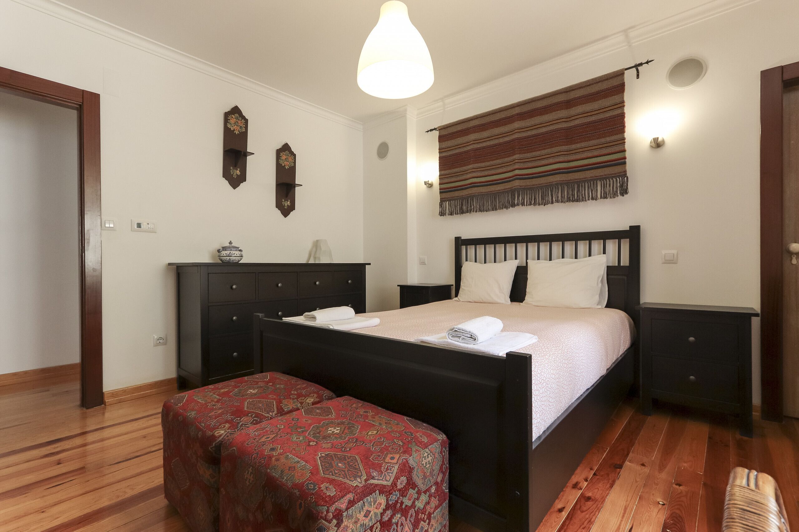 Une chambre avec un plancher en bois, un lit avec une tête de lit, une table de chevet, une commode, une horloge murale et une tapisserie suspendue.