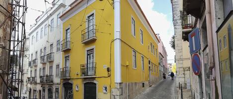 A narrow street with yellow and white buildings, parked cars, and a pedestrian in the distance.