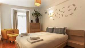 Un dormitorio moderno con una cama grande, un aparador de madera, una mesita pequeña, una silla y una lámpara de pared.