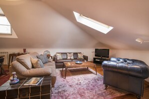An attic living room with a sloped ceiling, a sofa, a coffee table, a leather armchair, and a television.
