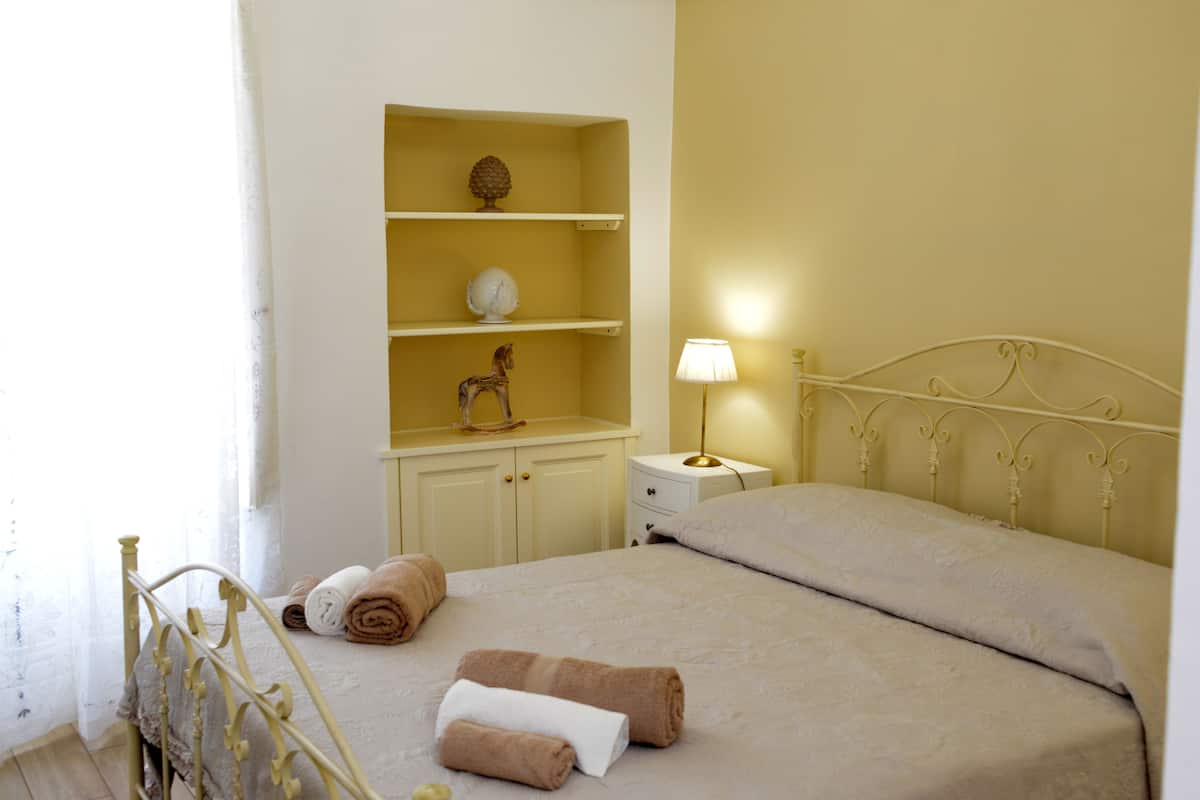 A neatly made bed with rolled towels, a bedside lamp, and a shelf with decorative items.