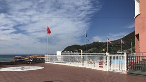 Um deck pavimentado com cerca branca, um mastro com bandeiras e vista para o mar e colinas ao fundo.