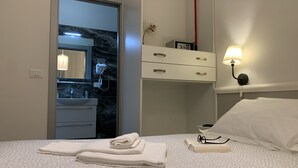Quarto de hotel com cama, mesa de cabeceira com luminária, espelho, guarda-roupa e banheiro visível através de uma porta aberta.