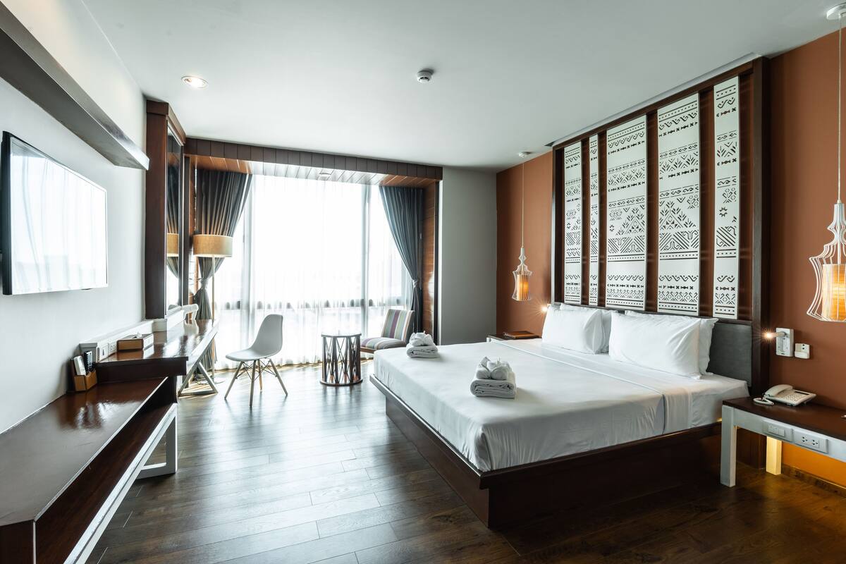 A modern hotel room with a large bed, a desk, a chair, and a view of the city through the window.