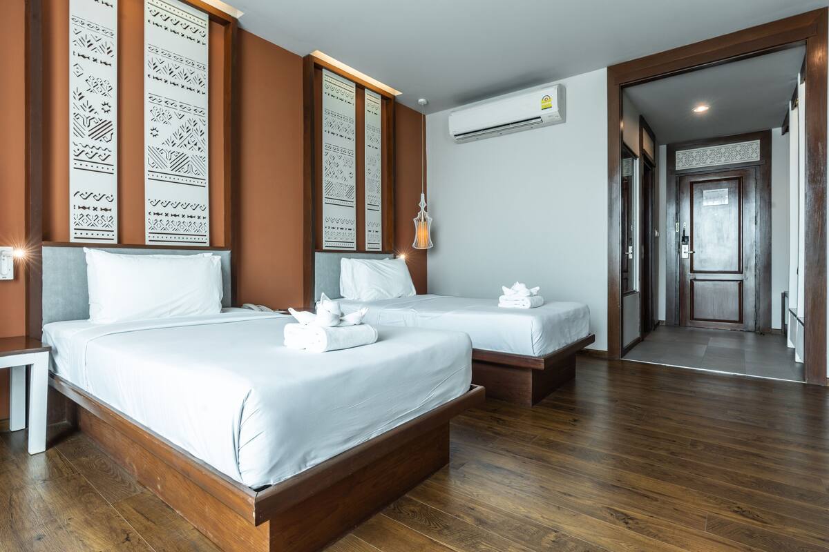 A hotel room with two beds, wooden flooring, a white wall with decorative panels, and an air conditioning unit.
