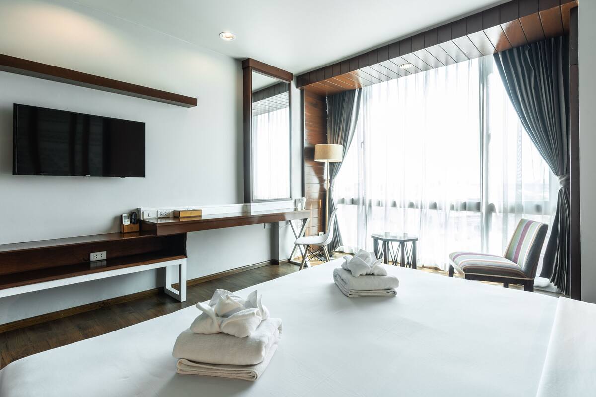 A hotel room with a large bed, a flat-screen TV mounted on the wall, a desk with a lamp, and a view of the city through large windows.