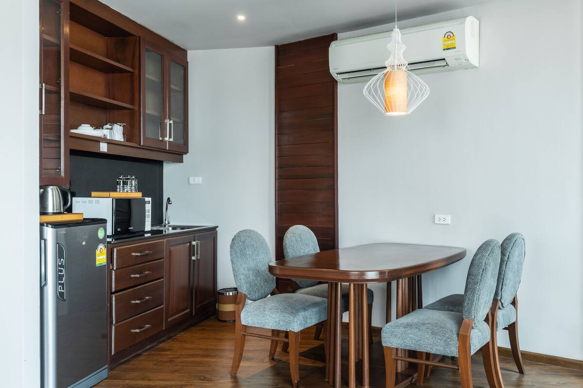 A compact kitchen with wooden cabinets, a stainless steel refrigerator, and a microwave. There is a dining table with four chairs and a hanging pendant light.