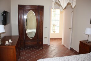 A bedroom with a wooden wardrobe, a bed with a floral bedspread, a bedside table with a lamp, and a television mounted on the wall. - B&B La Marchesa (Sant'Anastasia)