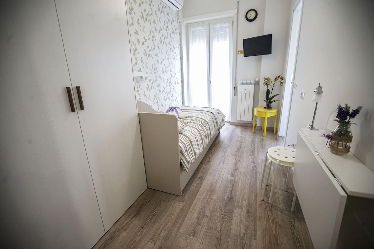 Un dormitorio con cama, armario, una mesita con una planta y televisor montado en la pared.
