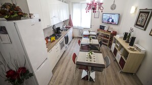 Cocina compacta con muebles blancos, una mesa de comedor con sillas, un refrigerador y un televisor en la pared.