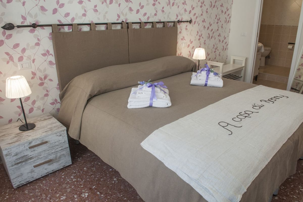 Habitación de hotel con cama, dos lámparas de noche, una mesita de noche con cajón y un baño visible a través de una puerta abierta.