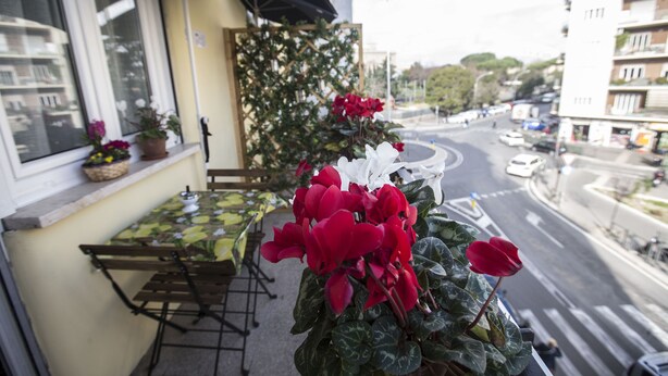 Un balcón con una mesa y sillas, una maceta con flores rojas, y vista a una calle de la ciudad.