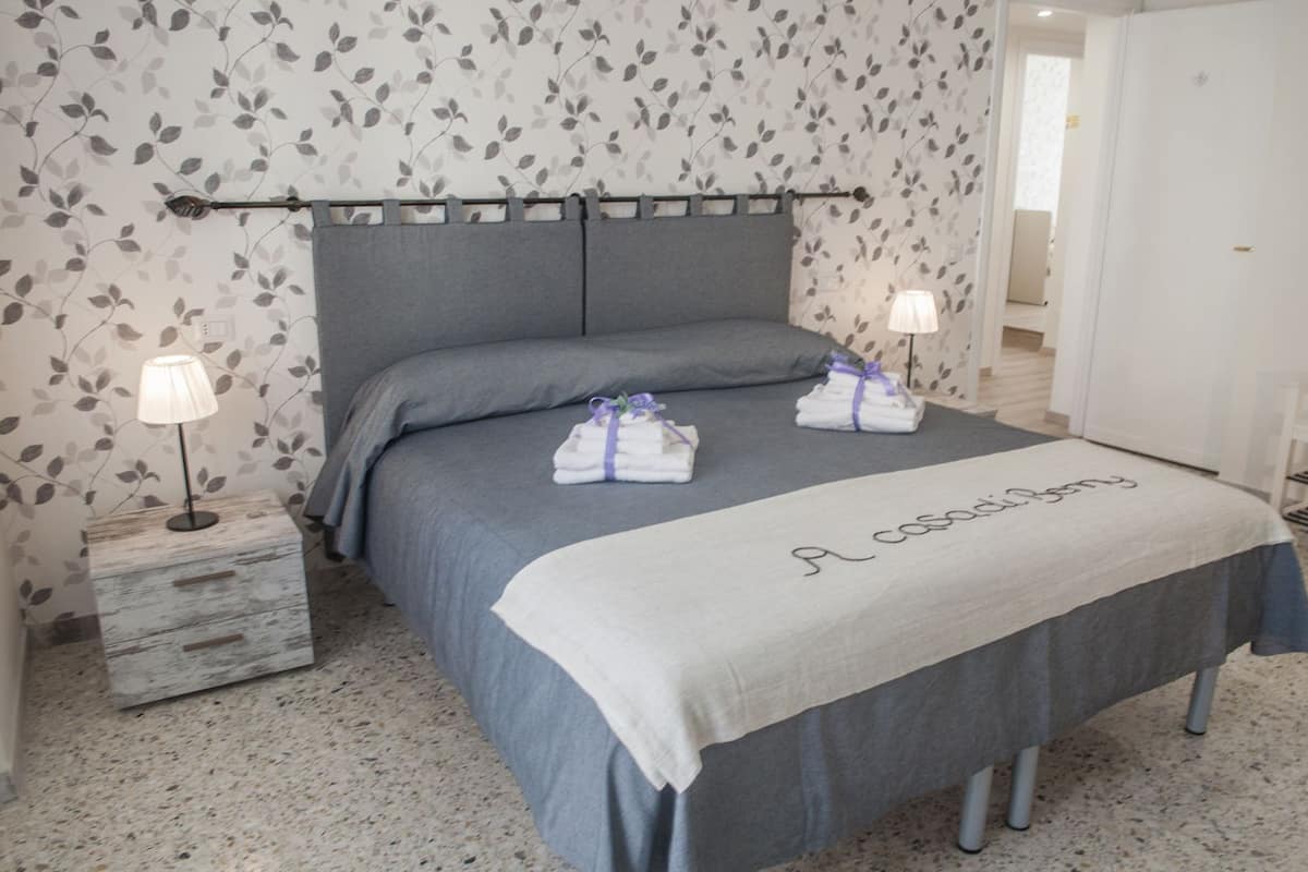 Una cama bien tendida con cabecera gris, dos lámparas de noche y una mesita de noche con lámpara. Se observa un perchero con toallas dobladas y un cartel sobre la cama.