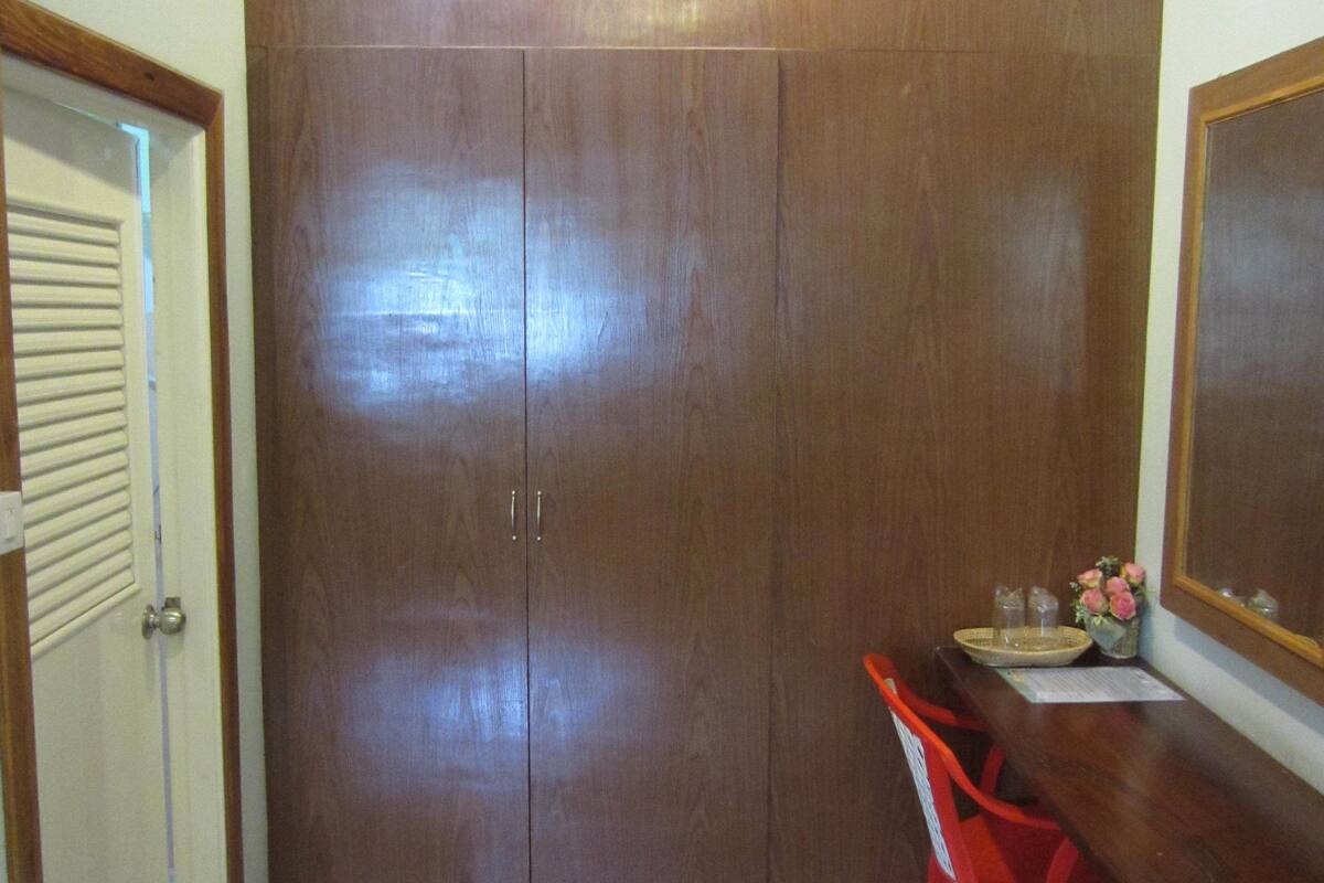 A room with a wooden wardrobe, a red plastic chair, a small table with a vase, and a mirror on the wall.