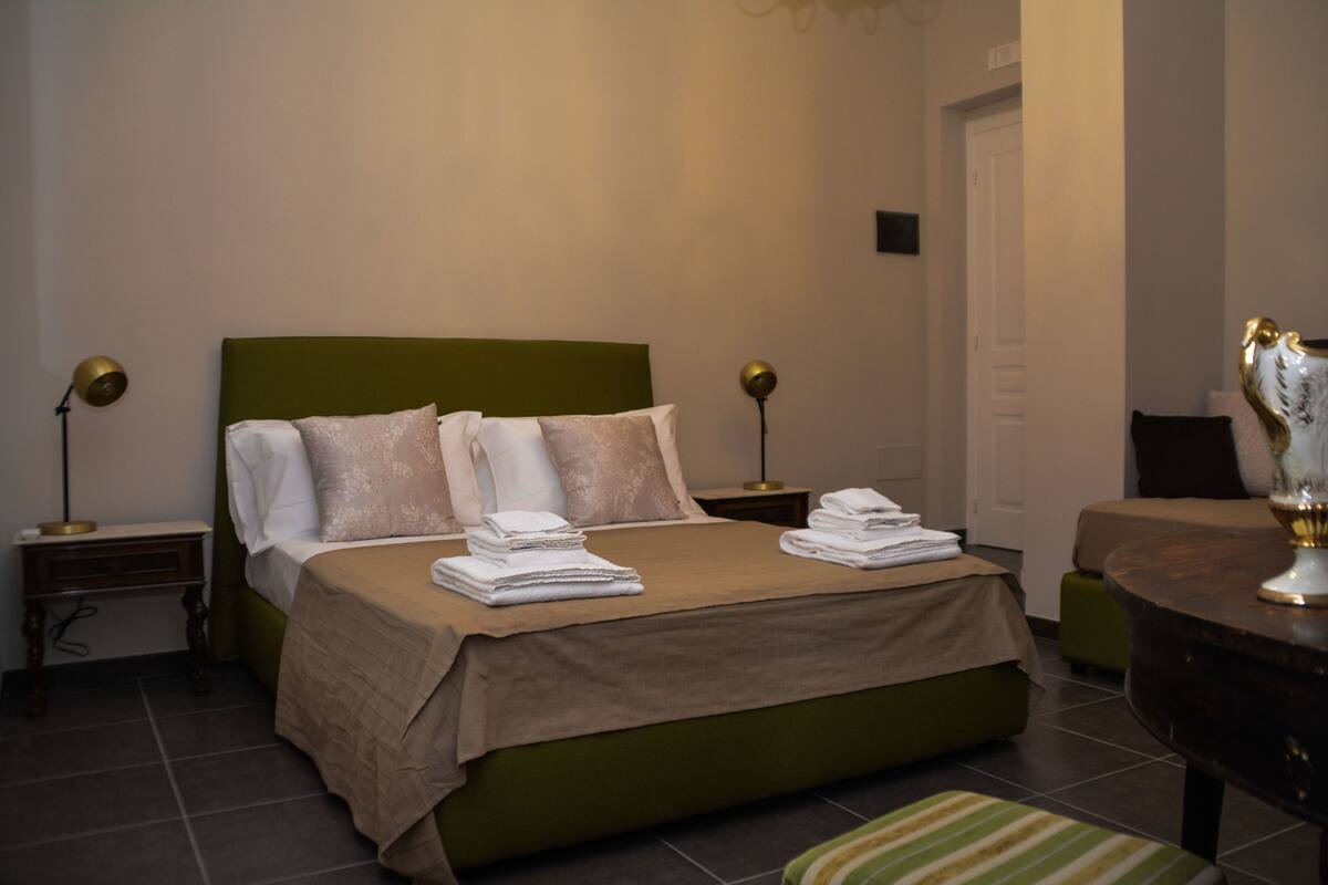 A neatly made bed with a green headboard, two bedside tables with lamps, and a small round table with a decorative item.