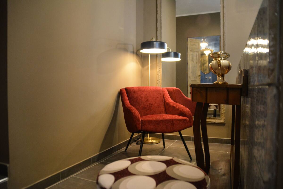 A red armchair, a round patterned rug, and a table with a lamp and a decorative item in a hallway with a mirror and a light fixture.