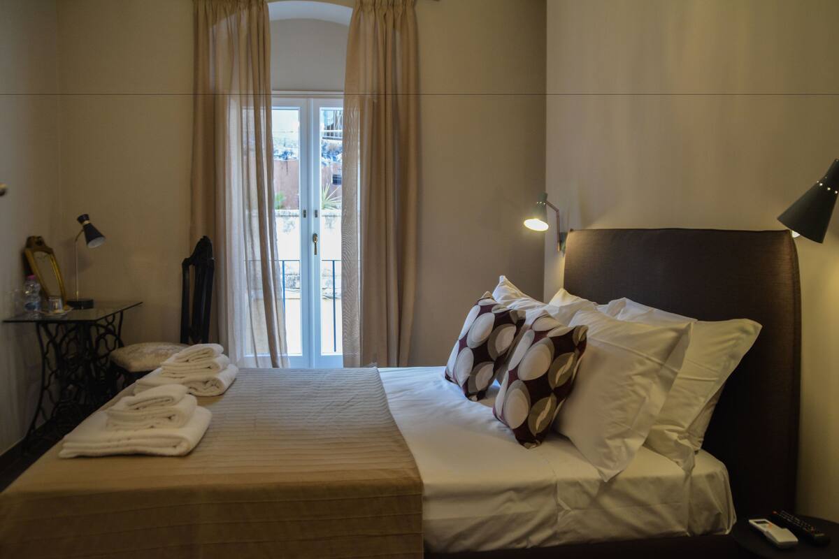 A hotel room with a bed, a table with towels, a lamp, and a view through a window with curtains.