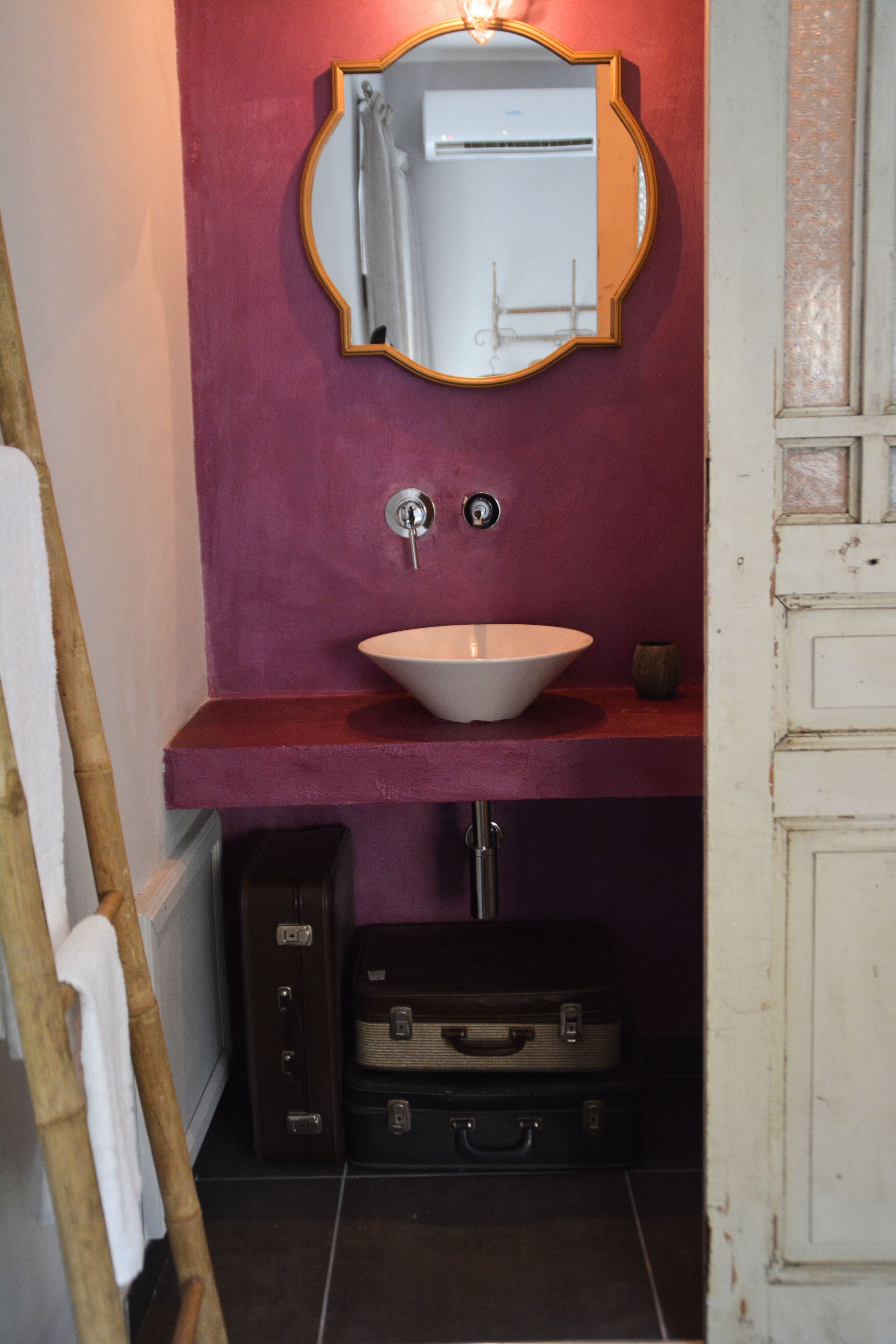 A bathroom with a white sink, a mirror, and a red wall. There is a suitcase under the sink.