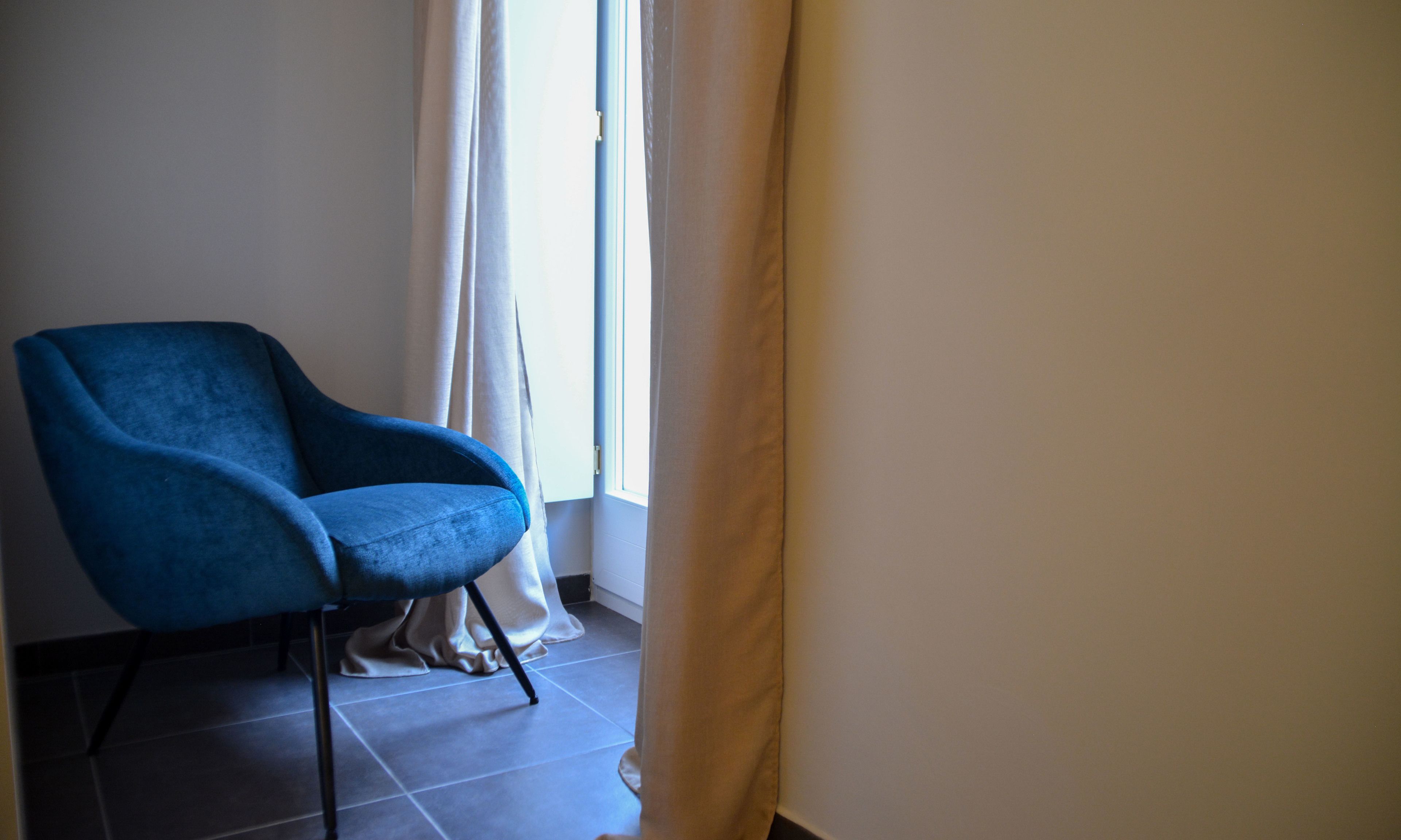 A room with a blue armchair, a window with white curtains, and a tiled floor.