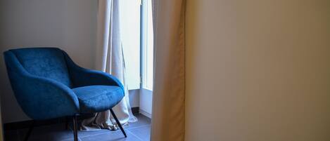 A room with a blue armchair, a window with white curtains, and a tiled floor.