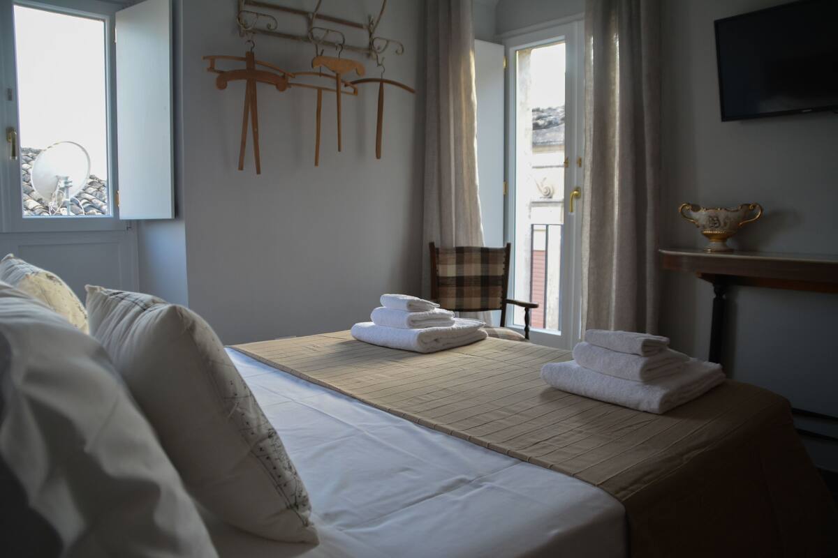 A hotel room with a bed, a table with towels, a chair, a television, and a window with curtains.
