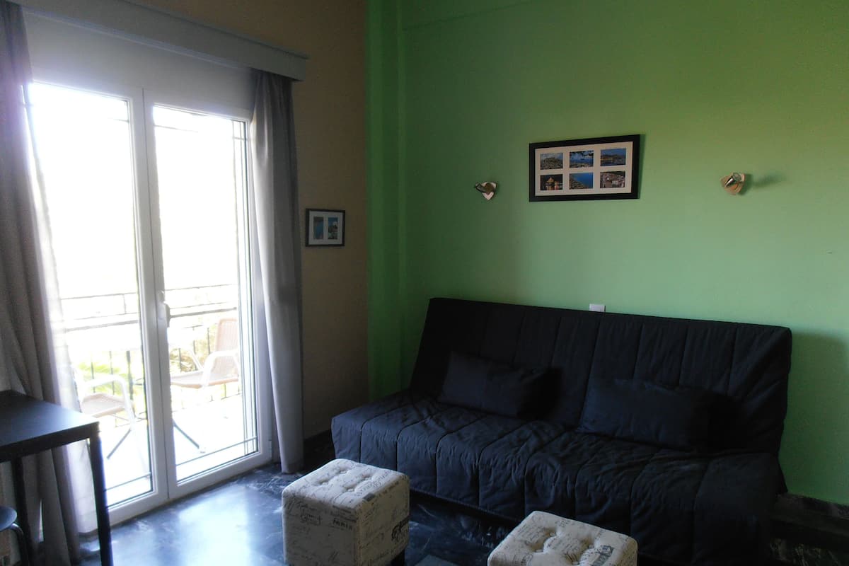 A room with a dark-colored sofa, a small table, and a sliding glass door leading to an outdoor area with chairs.