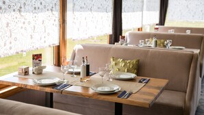 Comedor con mesas dispuestas para dos, que incluye una mesa de madera, sillas con tapicería color beige y vistas al exterior a través de cortinas de encaje.