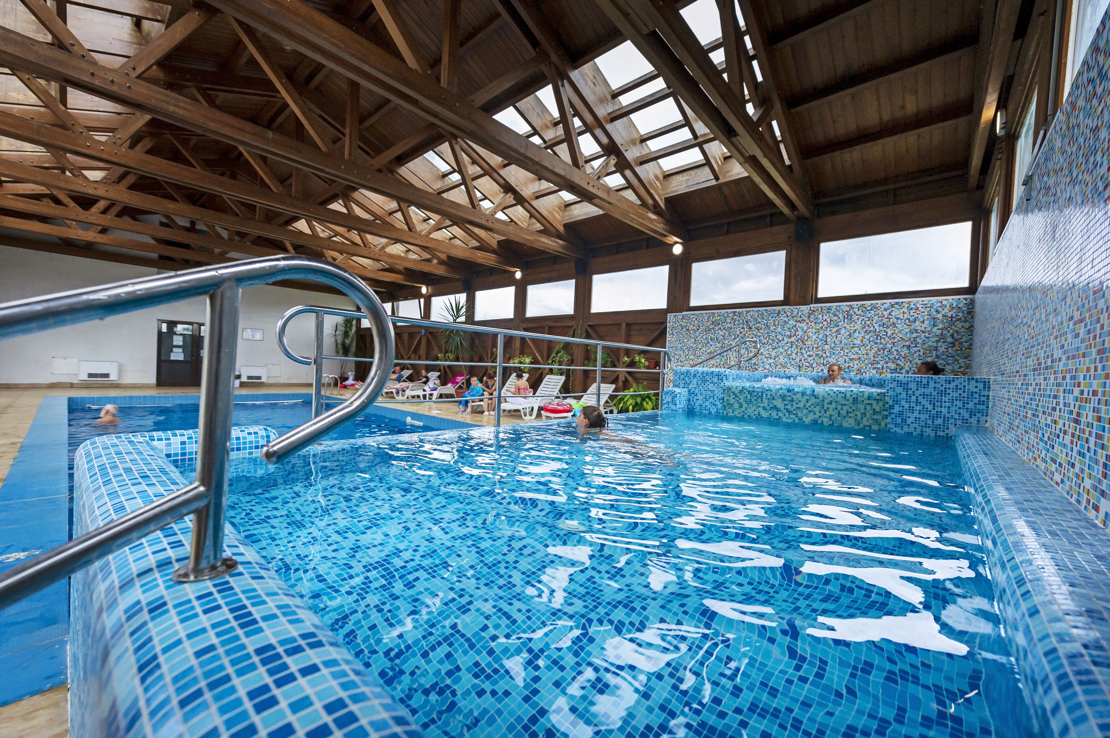 An indoor swimming pool with a tiled floor and walls, a metal handrail, and a wooden ceiling structure.