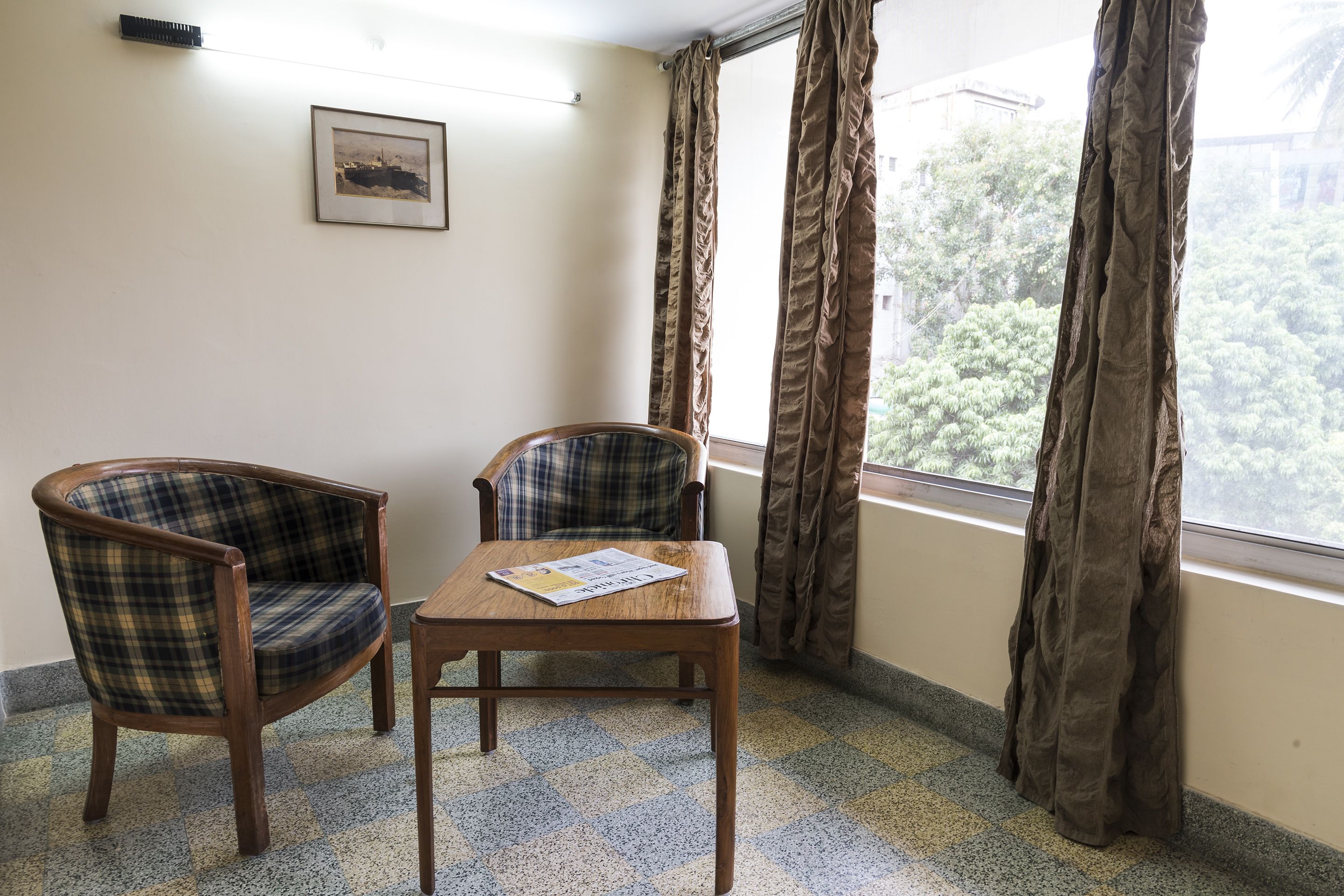 A room with two plaid chairs and a wooden table, a framed picture on the wall, and a window with curtains.