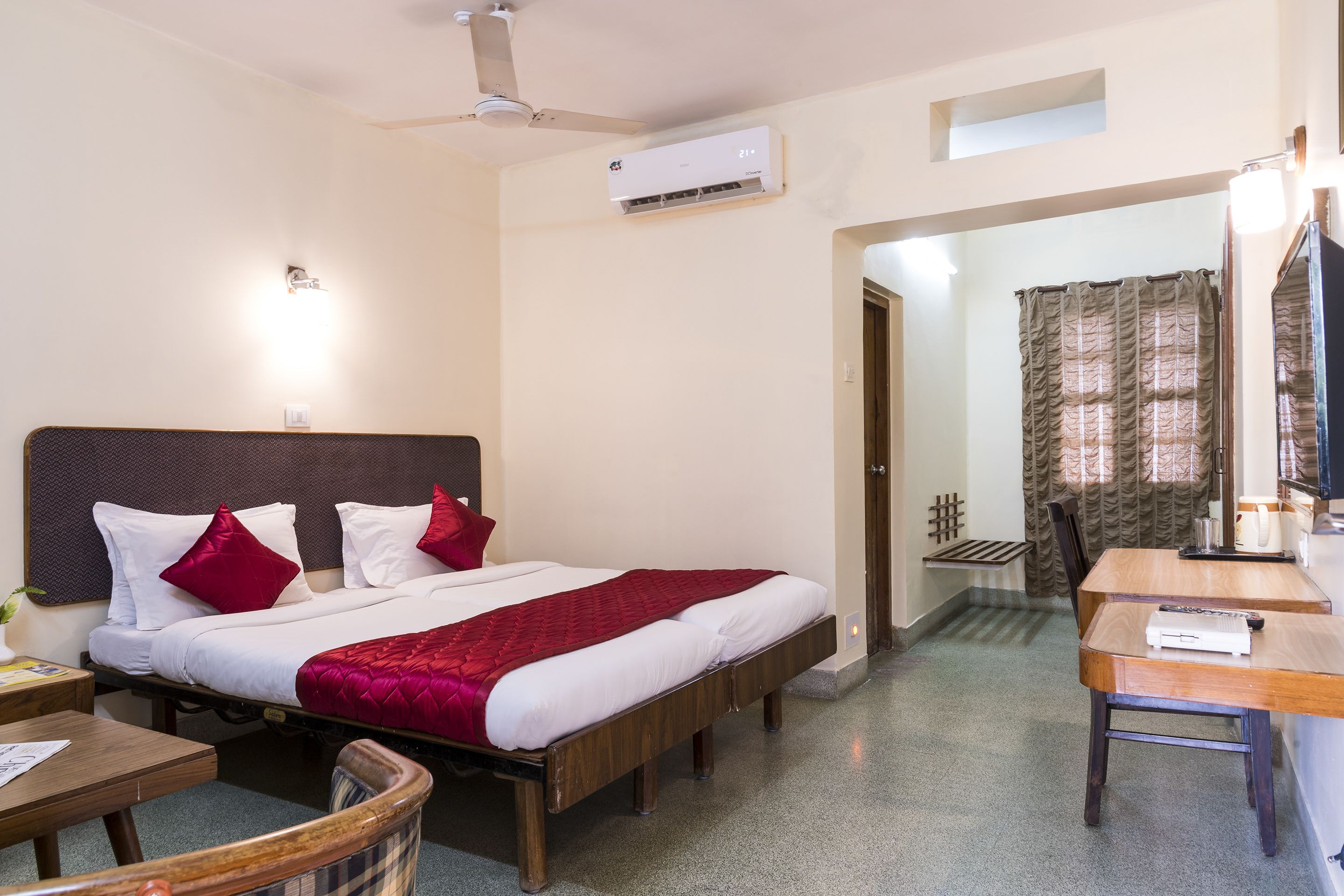 Habitación de hotel con una cama, un escritorio, una silla, un ventilador de techo, un equipo de aire acondicionado y un baño visible a través de una puerta abierta.