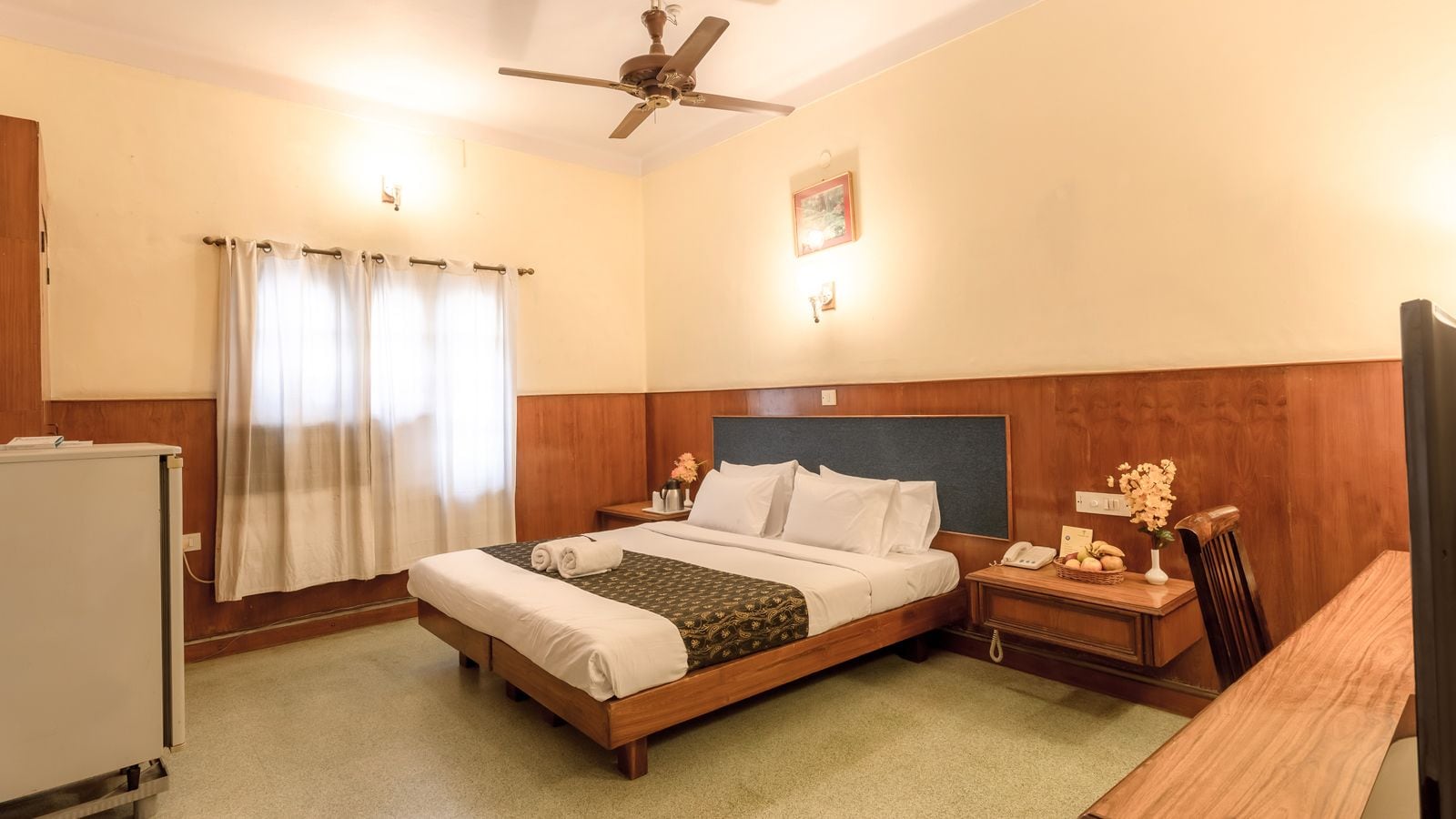 A hotel room with a bed, bedside tables, a desk, a chair, a ceiling fan, and a window with curtains.