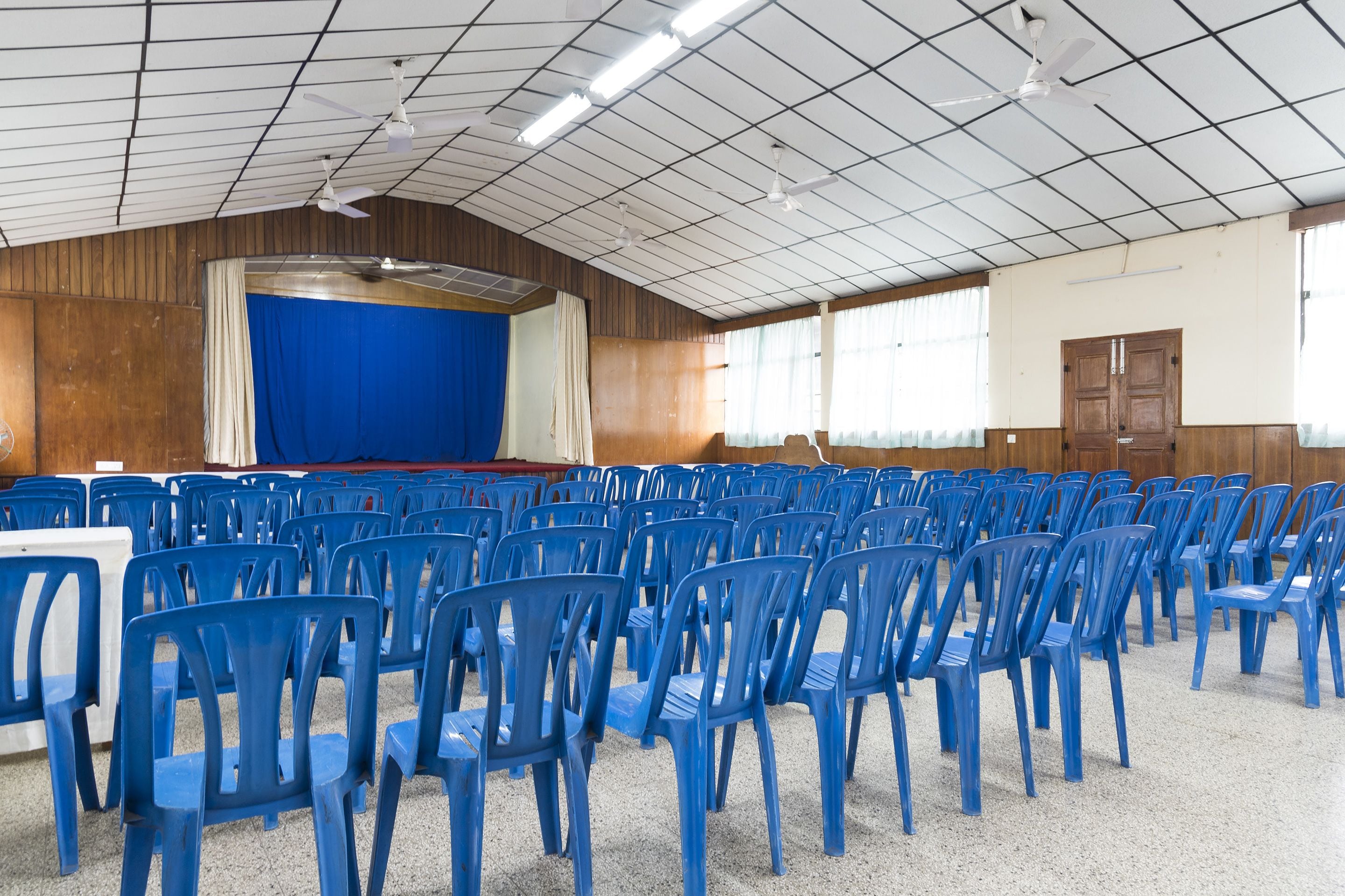 A large hall with rows of blue plastic chairs, a stage with a blue curtain, wooden panel walls, and a ceiling fan.
