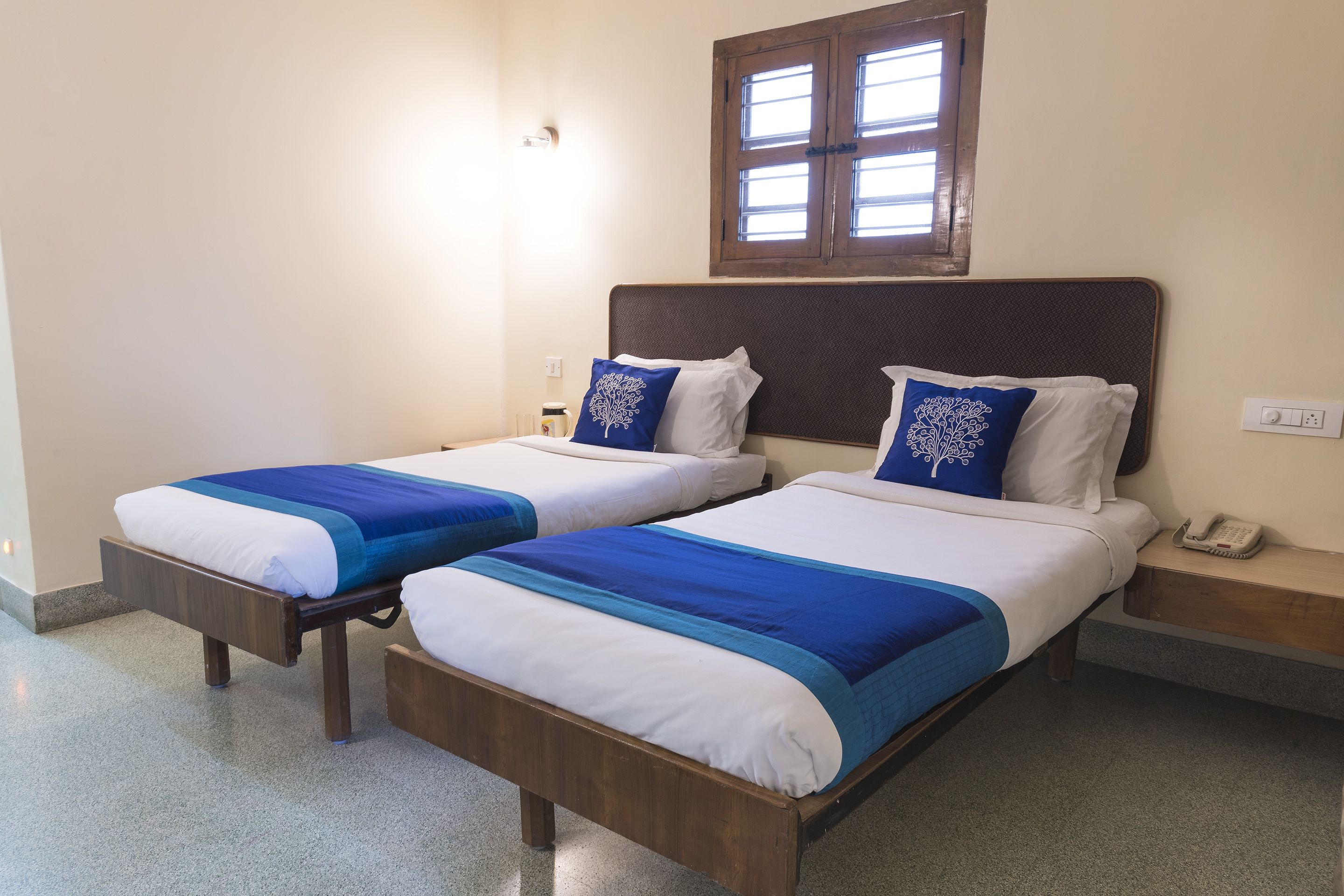 Habitación de hotel con dos camas individuales, cada una con colcha y almohadas azules y blancas. Tiene cabecera de madera, una mesita de noche con teléfono y una ventana con contraventanas de madera.