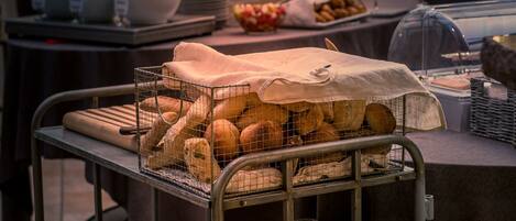 Un bufé con distintos alimentos, entre ellos pan y pasteles, expuestos en estantes metálicos.