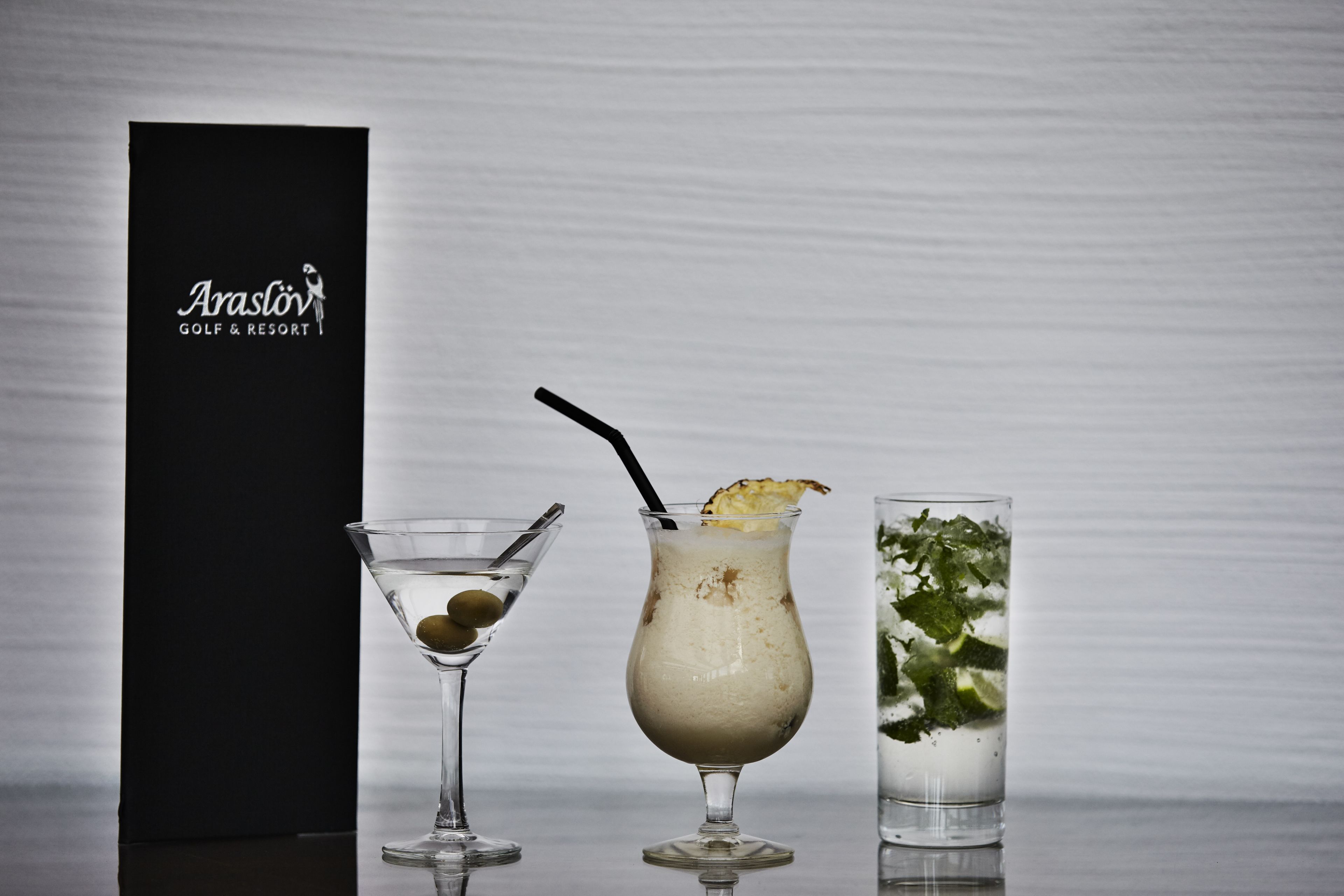 Tres bebidas sobre una mesa, con una caja negra que dice "Araslon Golf & Resort" al fondo.