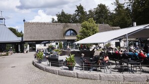 Ein Sitzbereich im Freien mit Tischen und Stühlen, ein Gebäude mit Strohdach und eine Steinmauer im Hintergrund.