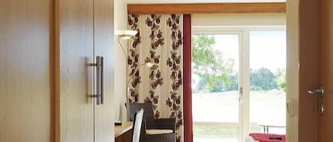 A room with a wooden wardrobe, a desk with a chair, a red sofa, and a large sliding glass door leading to an outdoor area.