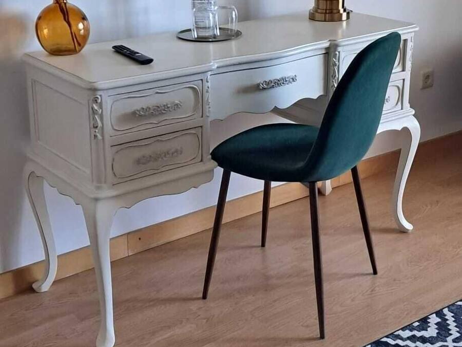Un escritorio blanco con una silla azul verdosa, una lámpara y un florero con plantas secas. En la pared, encima del escritorio, hay cuadros enmarcados.